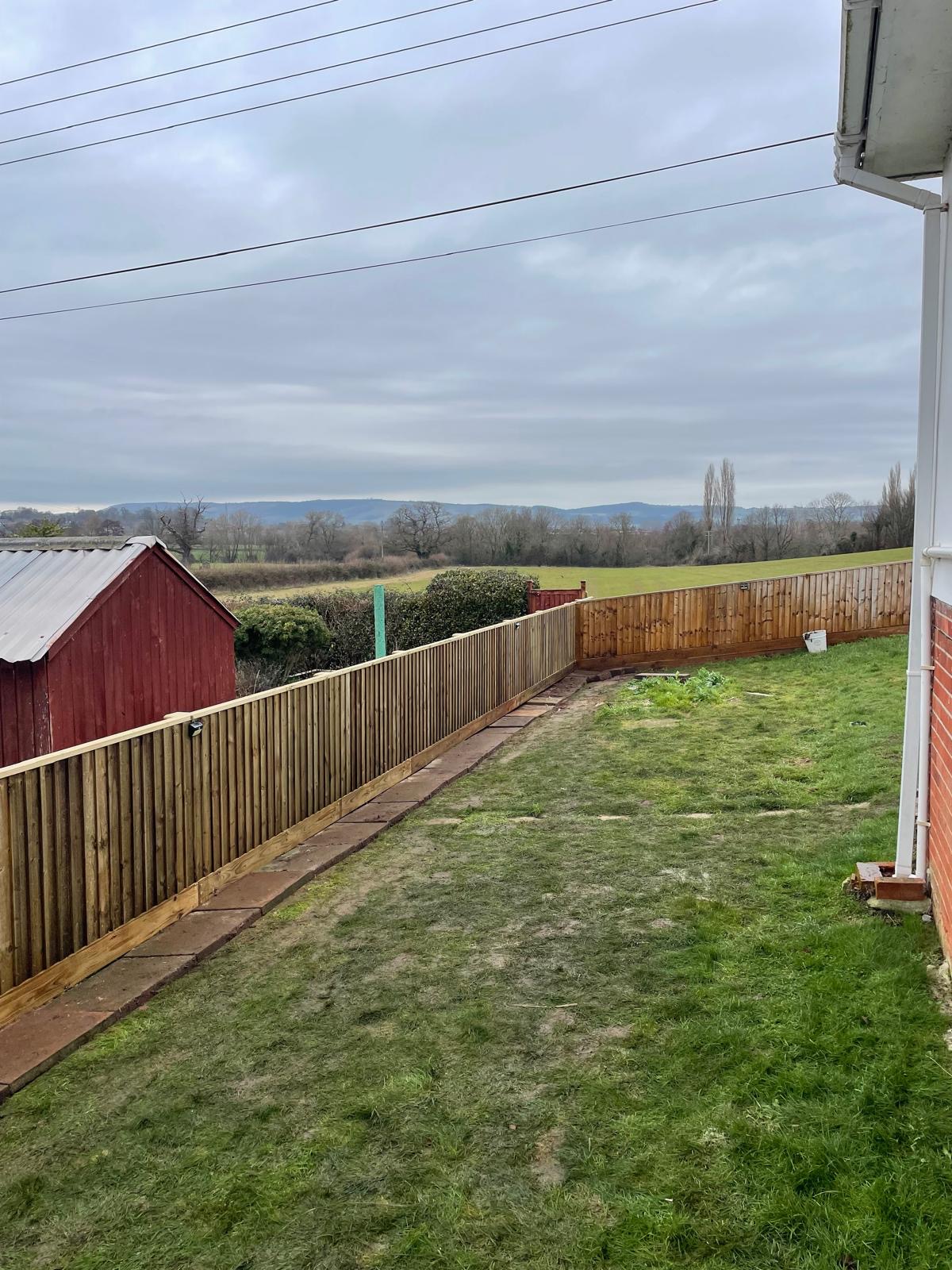 A long closeboard fence beside a lawn with fields and hills beyond